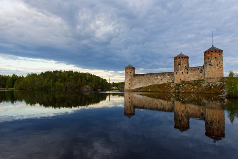 Olavinlinna's three stone towers rising from a rocky islet, reflected in the still lake water